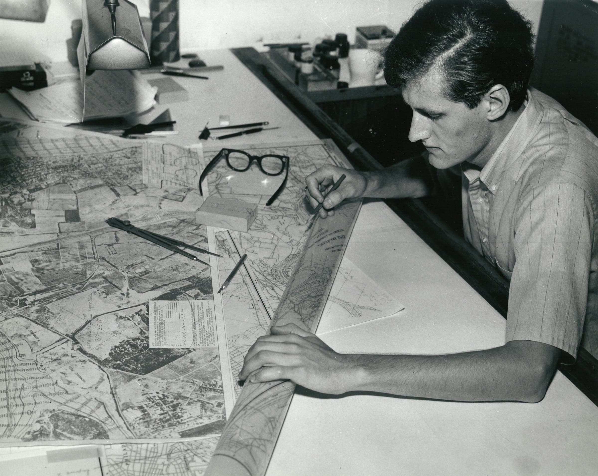 A man drawing on maps at a drafting table, in black and white