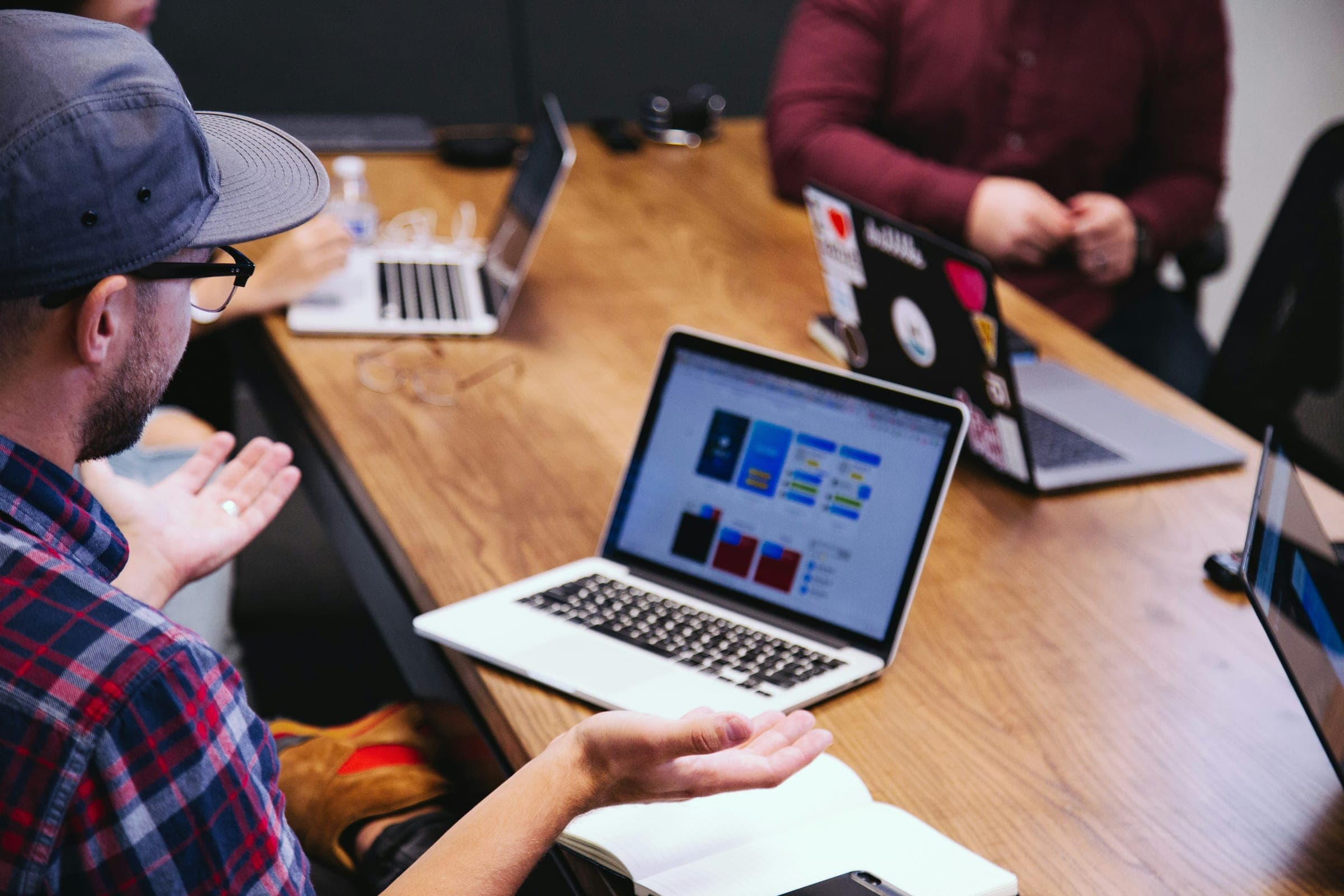 A man in a cap gesturing at a laptop with app designs during a team meeting