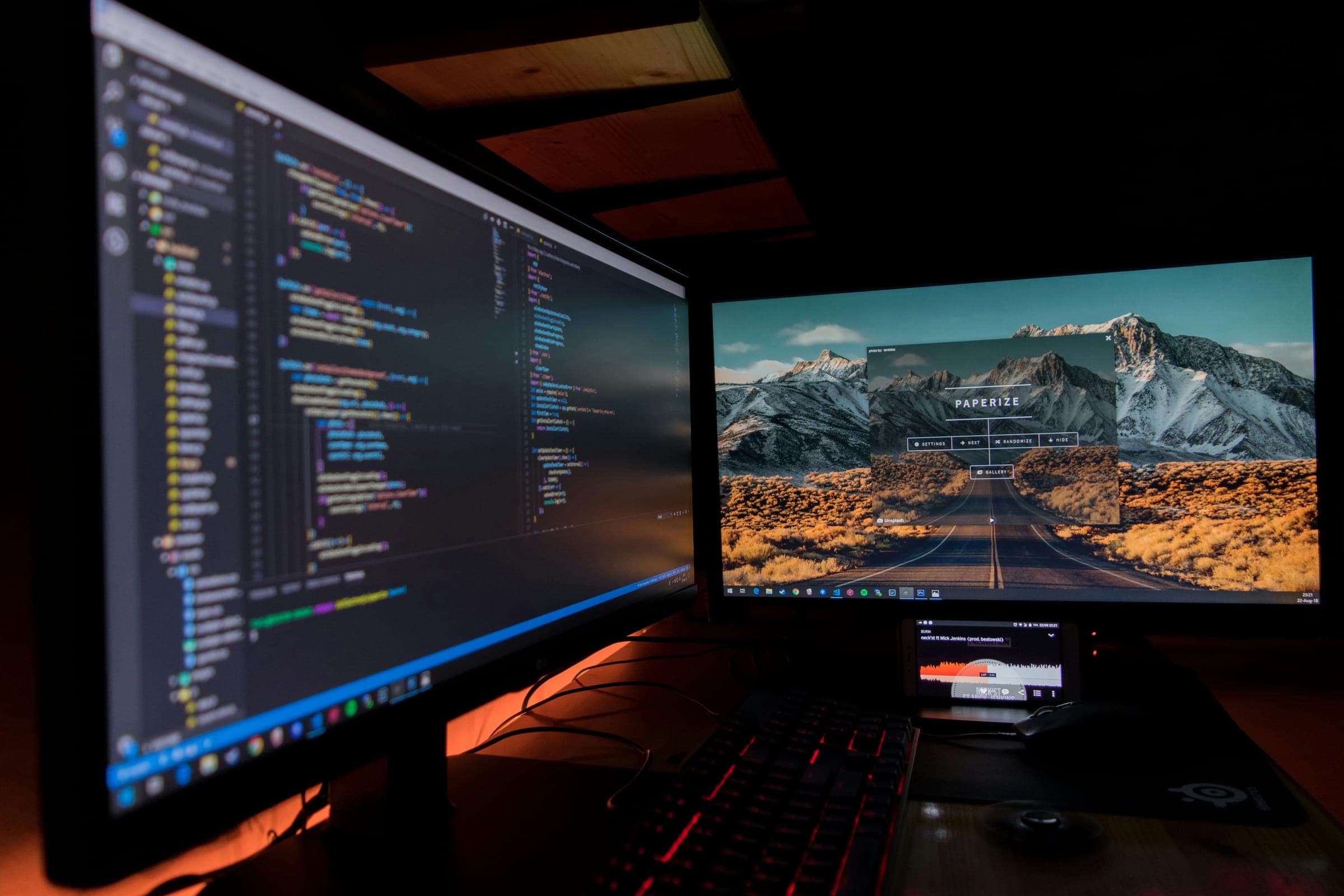Two monitors in a dark room, one with code and one with a mountain road